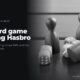 Board game pieces and dice on a wooden table, in black and white, beside the headline The card game carrying Hasbro